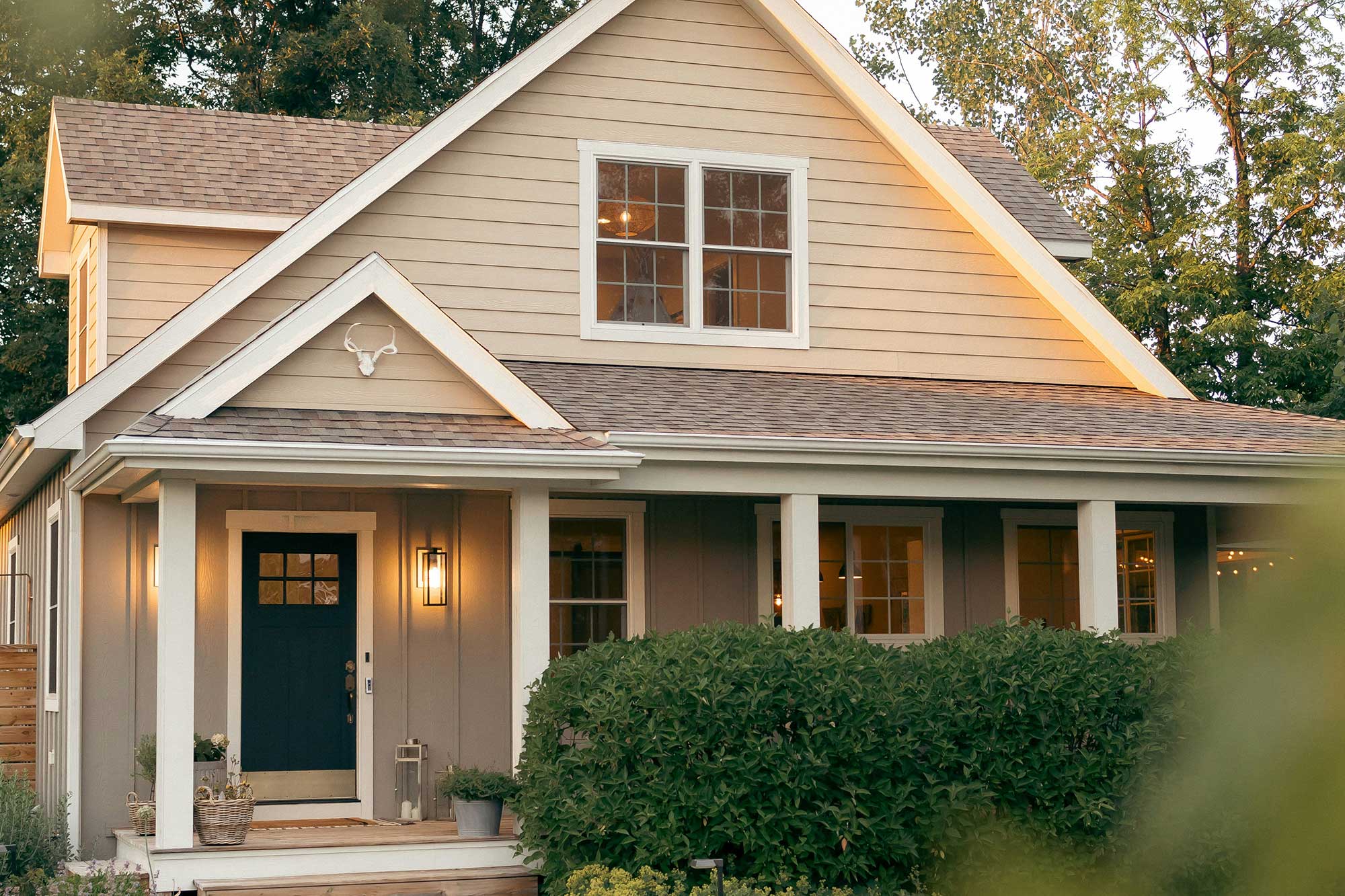 White 2-storey house with green door with indoor lights on and exterior porch light turn on, sitting behind green shrubs